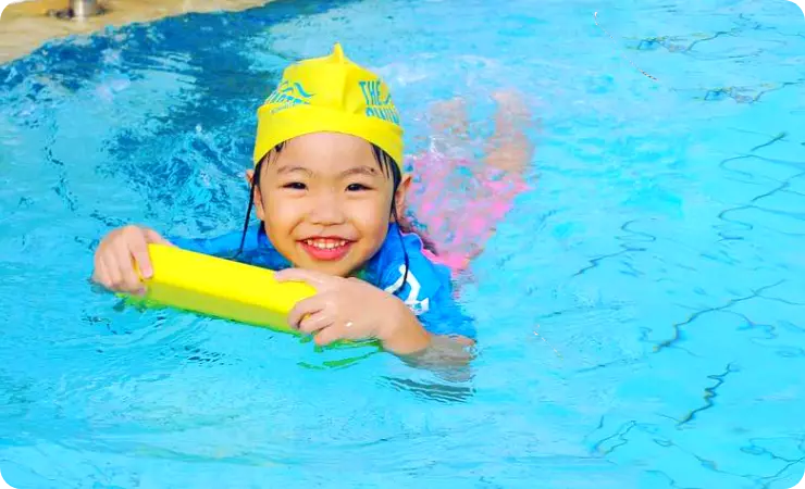 Kid wearing yellow swimming cap and learning how to swim in the deep pool on a yellow swimming board with no swimming experience during intermediate swimming lessons